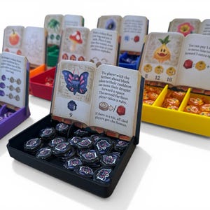 May include: A tabletop board game with various components. The game includes cards with illustrations, tokens, and containers in purple, red, yellow, and black. The cards feature images of a butterfly, a pineapple, and other game-related graphics.