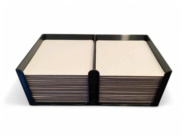 May include: A black card holder, rectangular in shape, divided into two sections, each containing a stack of white cards. The design is simple and modern, ideal for organising cards.
