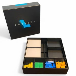 May include: A black box with the word "PROJECT" and a blue "L" logo. The box is open, revealing compartments with game pieces in yellow, green, blue, and orange. Two beige cards are also visible.