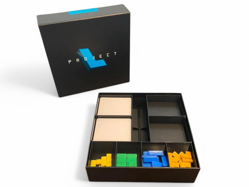 May include: A black box with the word "PROJECT" and a blue "L" logo. The box is open, revealing compartments with game pieces in yellow, green, blue, and orange. Two beige cards are also visible.
