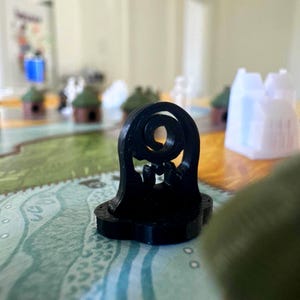 May include: A black, 3D-printed game piece with a stylized spiral design at the top. The base is a rounded rectangle. Other game pieces, including miniature houses, are visible in the background on a game board.