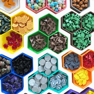 May include: A collection of board game pieces, including coins, wooden shapes, and dice, organized in colorful, hexagonal trays. The pieces are in various colors, such as yellow, green, red, and blue, and are arranged on a white surface.