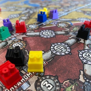 May include: A game board with various coloured plastic pieces, including red, green, yellow, black, and purple. The board features a map with the names "Kansas" and "Oklahoma" visible. The pieces are shaped like small buildings with pipes on top.