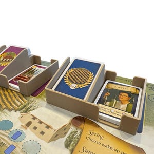 May include: A board game with card holders containing illustrated cards. The cards feature images of a winery, a gold wreath, and a person. The game board has a farm scene and the word "PLAY".