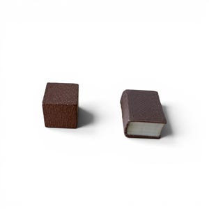 May include: Two miniature brown objects on a white background. One is a cube with a textured surface, and the other is a small book with a detailed cover and white pages. The book has a rounded spine.