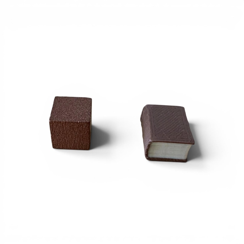 May include: Two miniature brown objects on a white background. One is a cube with a textured surface, and the other is a small book with a detailed cover and white pages. The book has a rounded spine.