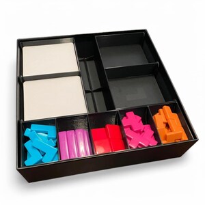 May include: A black board game organiser with several compartments. It contains white cards and colourful game pieces: blue, pink, red, and orange. The box is designed to store and sort board game components.
