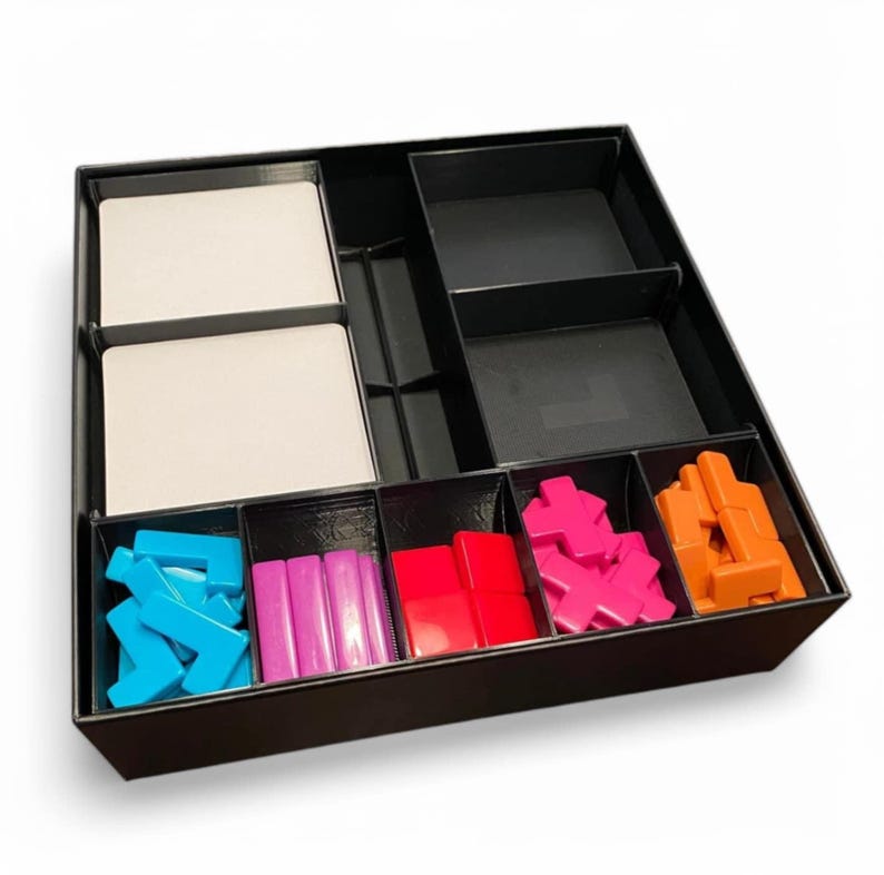 May include: A black board game organiser with several compartments. It contains white cards and colourful game pieces: blue, pink, red, and orange. The box is designed to store and sort board game components.