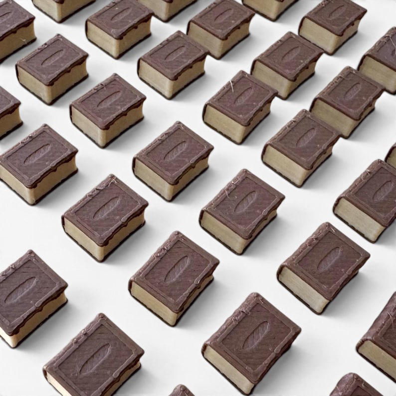 May include: A repeating pattern of miniature chocolate book-shaped candies. Each candy has a dark brown chocolate cover with a textured design and a lighter brown, rectangular base. The edges are lined with a darker chocolate.