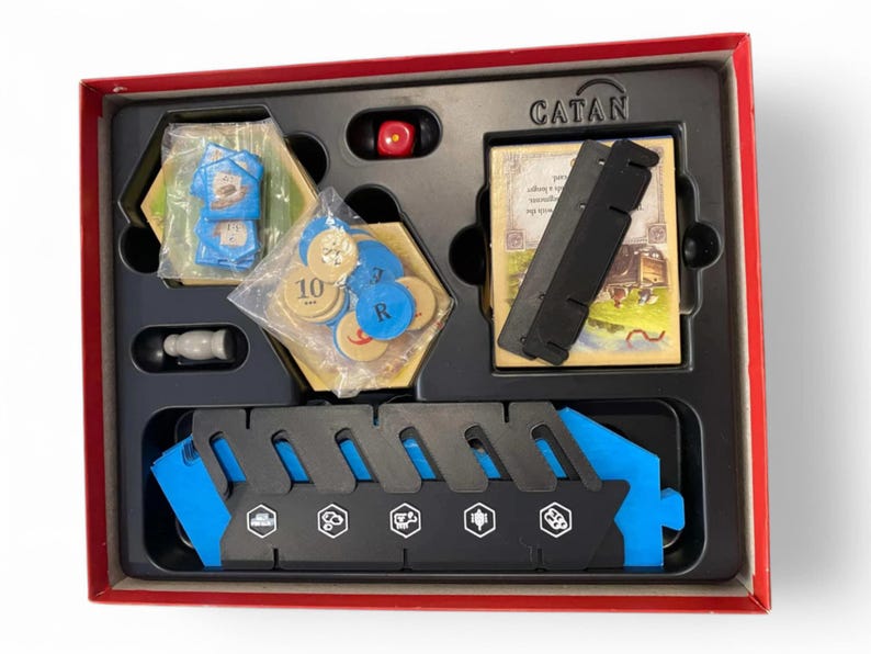 Puede incluir: Una caja roja con una bandeja negra que contiene piezas de juego de Catan. La bandeja tiene una barra de recursos azul y negra, un dado rojo y una ficha de madera con el n&uacute;mero 10. La caja tiene la palabra "CATAN" impresa.