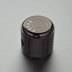 Puede incluir: Un botón de guitarra gris oscuro con un agarre texturizado con patrón de diamantes. La parte superior presenta un diseño circular con una carita sonriente y flechas. El botón es de metal y tiene una superficie brillante y reflectante.