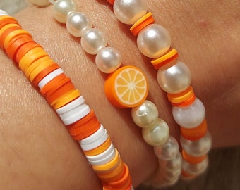 Op de afbeelding: Drie armbanden gestapeld op een pols. De onderste armband is gemaakt van oranje, witte en gele kralen. De middelste armband is gemaakt van witte parelkraaltjes met oranje en gele kralen. De bovenste armband is gemaakt van witte parelkraaltjes met oranje kralen en een enkele oranje schijf charme.
