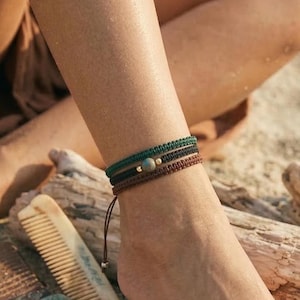 Può includere: Braccialetti alla caviglia nei toni del verde e del marrone con piccole perline. I braccialetti sono indossati su un piede appoggiato sulla sabbia. Un pettine, una cintura e una conchiglia sono anche sulla sabbia. Lo sfondo è sfocato, suggerendo un ambiente balneare.