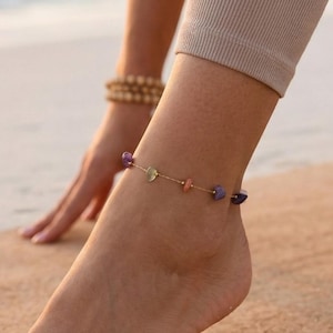 May include: A gold anklet featuring small, colourful gemstones. The anklet is worn on an ankle, with a light-coloured yoga mat and beach in the background. The person is wearing beige leggings.