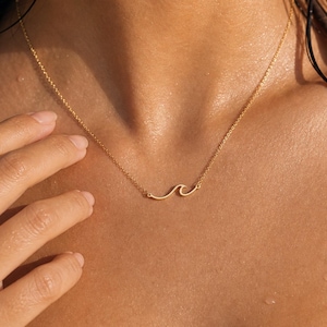 May include: A delicate gold necklace with a wave pendant. The necklace is worn on a person's chest, with the wave pendant resting near the collarbone. The gold chain is thin and the wave pendant is a simple, elegant design.