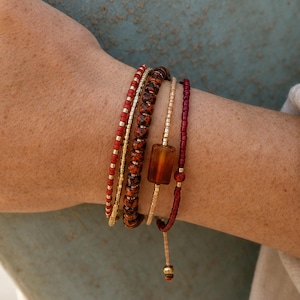 May include: A set of four stacked bracelets featuring red seed beads, a strand of dark brown faceted beads, a gold-toned chain with a rectangular amber-coloured bead, and a thin red cord with a small gold bead and a dangling end piece.