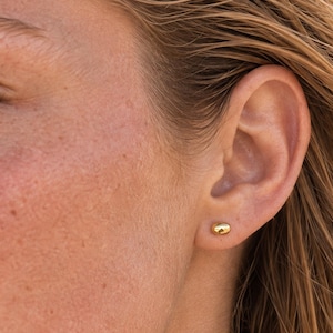 May include: A small, polished gold stud earring in a smooth, rounded pebble shape. The earring is worn in the earlobe, featuring a reflective metallic surface that catches the light. The design is simple, compact, and circular in appearance.