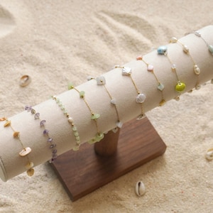 May include: A display stand holds several delicate bracelets. Each bracelet features a thin gold chain with various gemstone and pearl accents. The stand is made of wood and has a beige, cylindrical top. The background is a sandy surface.