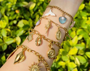 May include: Gold-toned charm bracelets featuring various ocean-themed charms. The bracelets include charms shaped like seashells, a sun, a mermaid tail, and a seahorse. The bracelets are layered and worn on a wrist against a backdrop of green foliage.