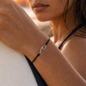 May include: A black cord bracelet with a silver wave charm. The bracelet is worn on a person's wrist, with a surfboard in the background. The image is taken outdoors during the golden hour.