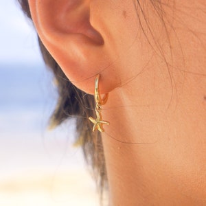 May include: Gold hoop earrings with a dangling starfish charm. The earrings are worn in a person's ear.