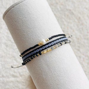 May include: A set of three layered bracelets featuring black cord, a single white pearl, a woven black and white patterned band, and a strand of small gold-toned beads interspersed with tiny grey faceted crystals on a cylindrical display.