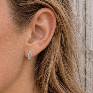 May include: A small, silver-coloured stud earring with a smooth, elongated oval shape. The earring is positioned securely in the earlobe, featuring a clean, metallic finish that reflects light evenly across its flat, rounded surface.