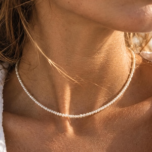 May include: A delicate choker necklace featuring a single strand of small, uniform, round white pearls. The necklace sits closely around the base of the neck, showcasing a simple and classic design with a smooth, lustrous finish on each bead.