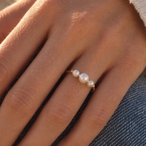 Può includere: Un sottile anello in oro con tre perle bianche di dimensioni diverse disposte in fila orizzontale sulla fascia. La perla centrale è la più grande, affiancata da due perle più piccole su entrambi i lati, creando un design delicato ed equilibrato.