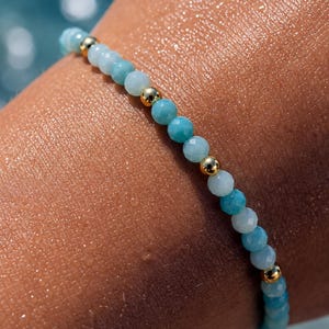 May include: A beaded bracelet featuring alternating small, faceted light blue and turquoise gemstone beads interspersed with smooth, polished gold-tone metal spherical beads, creating a delicate and uniform circular pattern around the wrist.