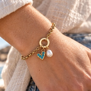 Könnte beinhalten: Ein goldfarbenes Gliederarmband mit einem runden Verbindungsring. An dem Ring hängen zwei Anhänger: ein kleines herzförmiges Element, das mit leuchtend blauen Steinen besetzt ist, und eine einzelne weiße, oval geformte Perle.