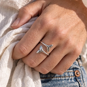 Op de afbeelding: Een zilverkleurige open ring met twee symmetrische walvisstaartmotieven die naar elkaar toe buigen. De gepolijste metalen band is verstelbaar, waarbij de twee uiteinden op de vinger zijn geplaatst om een open ontwerp te creëren.