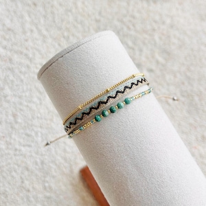 May include: A set of three layered bracelets featuring a thin gold-tone chain, a woven band with a black and white zigzag pattern, and a strand of small turquoise-coloured beads interspersed with tiny gold-tone metallic spacers and accents.