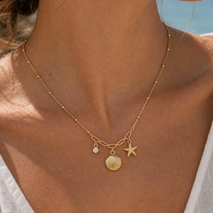 Peut inclure: Un collier doré composé d'une chaîne fine ornée de petites perles. La partie centrale comporte un petit cristal rond transparent, une breloque en forme de coquillage texturé et une étoile de mer, suspendus à un maillon ovale.