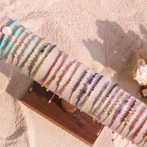 Könnte beinhalten: Eine Präsentation von Armbändern mit Perlen in verschiedenen Farben, darunter Türkis, Grün, Rosa, Lila und Rot. Die Armbänder sind auf einer hellfarbigen Schnur aufgereiht und weisen kleine, farbige Perlen auf. Sie sind auf einem grauen, zylindrischen Präsentationsständer mit Holzsockel angeordnet.