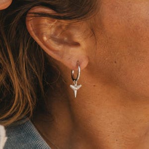 Peut inclure: Une boucle d'oreille créole en argent avec une petite breloque en forme de dent de requin. Le bijou est fabriqué dans un matériau métallique poli et pend au lobe de l'oreille, présentant un design simple avec un pendentif triangulaire.