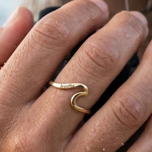 Könnte beinhalten: Ein goldfarbener Ring in Wellenform mit dem Wort "breathe" auf der Oberseite eingraviert. Der Ring wird an einem Finger getragen, mit einem verschwommenen Ozeanhintergrund.