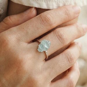 May include: A silver ring featuring a light blue, raw-cut gemstone. The ring is displayed on a hand, showcasing the natural, irregular shape of the stone. The setting appears simple, highlighting the gemstone's color and texture.