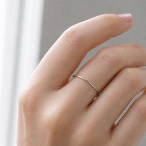 May include: A thin, smooth, silver-colored metal ring with a simple, circular band design. The ring features a polished, reflective surface that catches the light, creating a subtle metallic sheen. It is a minimalist piece of jewelry.