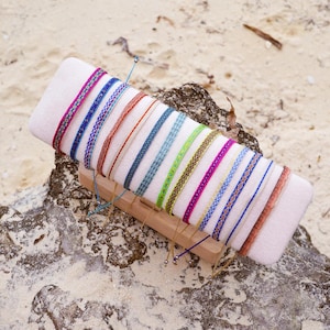 May include: A display of colorful woven bracelets on a white rectangular cushion. The bracelets feature various patterns and colors, including pink, blue, green, and orange. The display sits on a wooden base, resting on a rock and sand background.
