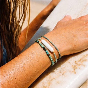 May include: Three beaded bracelets on a person's arm. One bracelet has a white, oblong bead. The other two bracelets are turquoise and gold. The person is holding a white surfboard.
