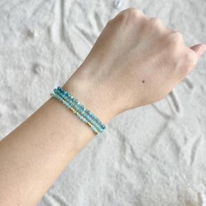 May include: A three-strand bracelet with small, light blue beads and gold-coloured accents. The bracelet is displayed on a wrist, highlighting the delicate design and colour variations of the beads. The beads are strung together, creating a layered effect.