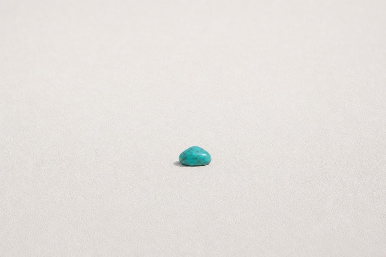 May include: A single, small, polished turquoise gemstone with a smooth, rounded, dome-like shape. The stone displays a vibrant cyan-blue color with subtle, darker natural veining patterns distributed across its polished, reflective surface.
