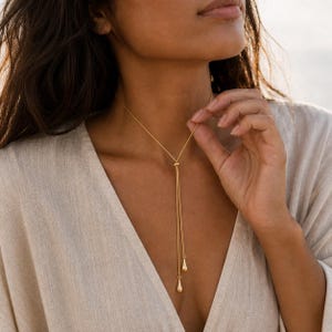 Puede incluir: Un collar tipo lazo en tono dorado que presenta una cadena fina que pasa a través de una pequeña cuenta deslizante, terminando en dos colgantes en forma de lágrima de diferentes longitudes. El collar descansa sobre el cuello y el pecho.