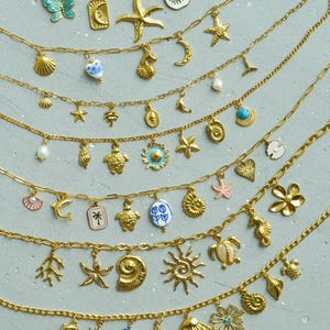 Könnte beinhalten: Eine Sammlung von goldenen Halsketten mit verschiedenen Anhängern, darunter Muscheln, Seesterne, Schildkröten, Blumen und andere nautische Designs. Die Halsketten sind auf einem grauen Hintergrund übereinander gelegt.