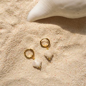 May include: Two gold-colored hoop earrings with white seashell charms, resting on a bed of sand. A large white seashell is in the background. The earrings have a simple, elegant design, perfect for beach-themed jewelry.
