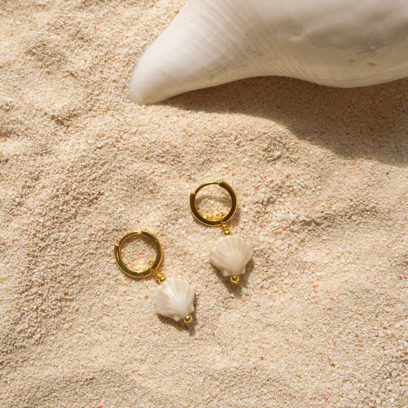 May include: Two gold-colored hoop earrings with white seashell charms, resting on a bed of sand. A large white seashell is in the background. The earrings have a simple, elegant design, perfect for beach-themed jewelry.