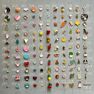 K&ouml;nnte beinhalten: Eine Sammlung von 100 Emaille-Anh&auml;ngern in verschiedenen Farben und Designs. Die Anh&auml;nger sind in Reihen auf einer grauen Oberfl&auml;che angeordnet. Die Anh&auml;nger umfassen Herzen, Sterne, Ananas, Eiswaffeln und mehr.