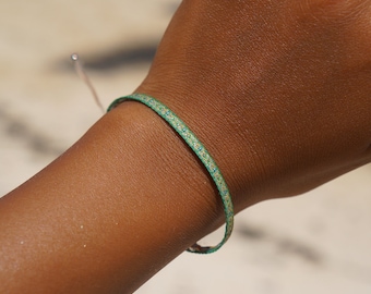 May include: A close-up of a green and blue bracelet on a wrist. The bracelet is thin and appears to be made of woven material. The background is blurred, suggesting an outdoor setting. The bracelet is a simple, minimalist accessory.