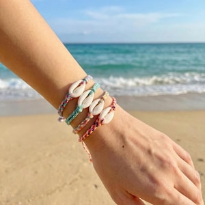 May include: Four colourful woven bracelets, each with a white seashell, are shown on a wrist. The bracelets are in shades of pink, blue, and green. The background shows a beach with the ocean in the distance.